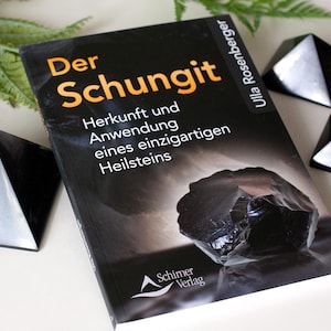 Könnte beinhalten: Ein Buch mit dem Titel "Der Schungit" mit schwarzem und orangefarbenem Text, das eine dunkle Mineralillustration zeigt. Drei schwarze pyramidenförmige Steine sind um das Buch auf einer weißen Oberfläche angeordnet, mit grünem Laub im Hintergrund.