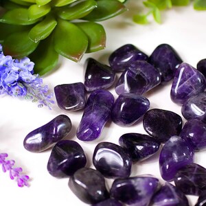 Könnte beinhalten: Eine Sammlung glatter, polierter Amethyststeine in verschiedenen Violetttönen. Die Steine sind auf einer weißen Oberfläche angeordnet.
