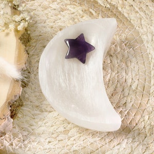 Könnte beinhalten: Eine weiße, mondsichelförmige Schale aus Selenit mit einer glatten, faserigen Textur. In der Schale liegt ein kleiner, polierter, fünfzackiger Stern aus tiefviolettem Amethyst-Edelstein mit einer glänzenden, reflektierenden Oberfläche.