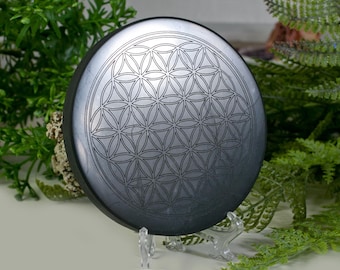 May include: A circular, dark grey stone featuring an etched Flower of Life design. The stone is presented on a clear stand, with green plants in the background. The pattern consists of interconnected circles.