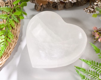 May include: A white, heart-shaped selenite bowl. The bowl has a smooth, opaque surface with slight textural variations. The image is styled with green foliage and a woven basket, creating a natural aesthetic.
