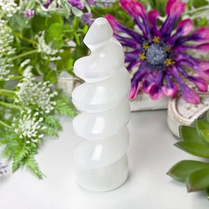 Könnte beinhalten: Eine weiße Selenitturm-Skulptur mit einem Spiraldesign. Die Skulptur ist von Grün und lila Blumen umgeben, was eine natürliche und dekorative Ästhetik schafft. Der Turm ist ein dekorativer Gegenstand.