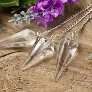 Può includere: Tre pendoli di cristallo di quarzo trasparente con catene d'argento sono appoggiati su una superficie di legno rustico. I pendoli sono sfaccettati e appuntiti, con un aspetto trasparente. Fiori viola e fogliame verde sono disposti intorno.
