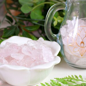 May include: A white ceramic bowl filled with pink rose quartz crystals. A glass pitcher with a gold lotus design is also filled with rose quartz crystals.