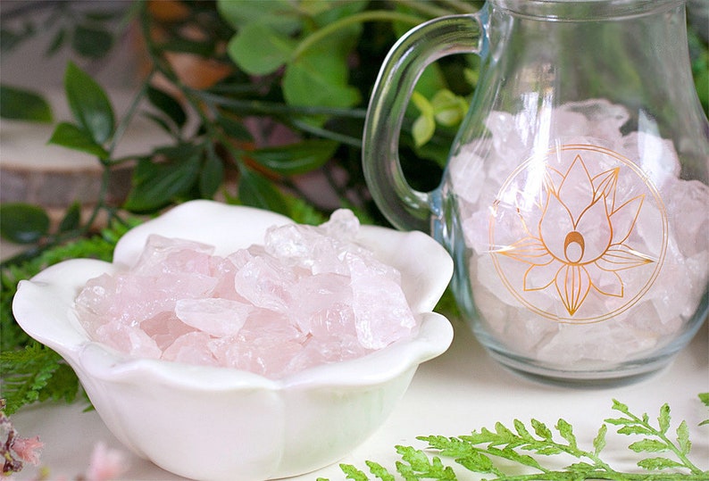 May include: A white ceramic bowl filled with pink rose quartz crystals. A glass pitcher with a gold lotus design is also filled with rose quartz crystals.