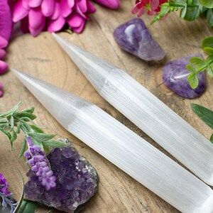 Könnte beinhalten: Zwei weiße Selenit-Stäbe, auch bekannt als Selenit-Sticks, auf einer Holzoberfläche mit violetten Amethyst-Kristallen und rosa Blüten. Die Stäbe sind glatt und haben eine gestreifte Textur.