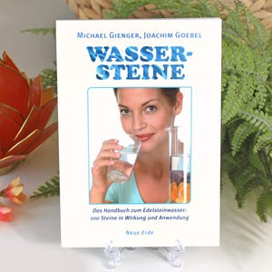 May include: A white book with a blue title that reads "WASSER-STEINE" and a subtitle that reads "Das Handbuch zum Edelsteinwasser: 100 Steine in Wirkung und Anwendung." The book is on a clear plastic stand and features a photo of a woman drinking from a glass.