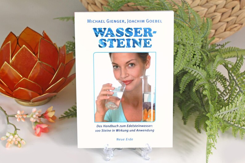 May include: A white book with a blue title that reads "WASSER-STEINE" and a subtitle that reads "Das Handbuch zum Edelsteinwasser: 100 Steine in Wirkung und Anwendung." The book is on a clear plastic stand and features a photo of a woman drinking from a glass.