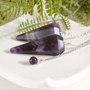 Op de afbeelding: Twee amethist kristallen pendels met zilveren kettingen liggen op een wit bord, met groen blad. De pendels zijn facetgeslepen, diep paars, met een kleine paarse kraal aan het einde van de ketting. De kettingen zijn zilver.