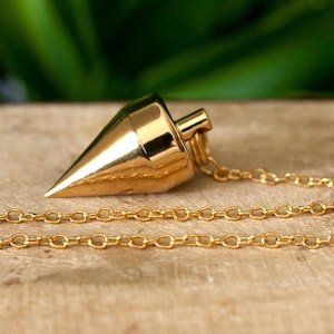Op de afbeelding: Een goudkleurige pendel met een conische vorm en een ketting. De ketting is bevestigd aan een kleine, ronde, witte parel. De pendel is ontworpen voor waarzeggerij en spirituele praktijken.