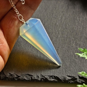 Op de afbeelding: Een facetgeslepen, opalescent kristallen pendel, opgehangen aan een fijne zilveren ketting. Het kristal heeft een lichtblauwe en witte kleur, met een puntige punt. De pendel ligt op een donkergrijze ondergrond.