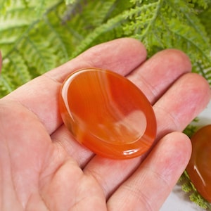 May include: Two orange carnelian palm stones, one in a hand and one on a white surface. The stones are smooth and polished with visible banding.