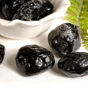 Könnte beinhalten: Mehrere polierte schwarze Steine, möglicherweise Obsidian oder ein ähnliches Material, werden gezeigt. Einige befinden sich in einer weißen, geriffelten Schale, andere sind auf einer weißen Oberfläche verstreut. Ein Zweig grünes Laub und eine kleine orangefarbene Blume sind im Hintergrund.