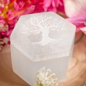 Könnte beinhalten: Weiße Selenit-Hexagon-Box mit einem eingravierten Baum des Lebens auf der Oberseite. Die Box steht auf einer Holzoberfläche mit rosa Blumen im Hintergrund.