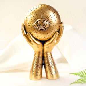 Può includere: Una scultura decorativa color oro con un design stilizzato a occhio su un disco rotondo, tenuto da due mani. Le mani e il disco sono adornati con motivi e dettagli intricati. La scultura è ambientata su uno sfondo bianco morbido.