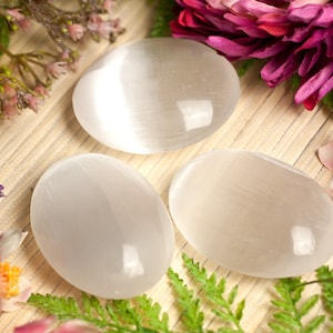 Può includere: Tre pietre lisce di selenite bianca, note anche come spato satinato, sono disposte su una superficie di legno marrone chiaro. Le pietre hanno una forma ovale e hanno un leggero luccichio. Lo sfondo presenta fiori rosa e viola.