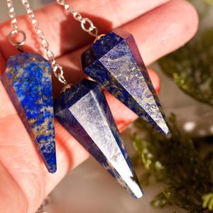 Può includere: Tre pendenti in lapislazzuli sfaccettati, ciascuno con una punta e una base esagonale, sospesi a catene in metallo color argento. Le pietre sono di un blu intenso con inclusioni di pirite color oro visibili sulle superfici levigate.