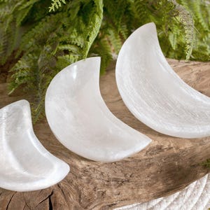 Può includere: Tre ciotole a forma di mezzaluna intagliate in cristallo di selenite bianco traslucido. Le ciotole sono disposte in fila, ognuna caratterizzata da una superficie liscia e lucida con una consistenza fibrosa e madreperlacea tipica della pietra.