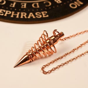 Pode incluir: Um pêndulo cor de cobre com um design em espiral e uma ponta, ligado a uma corrente. O pêndulo está sobre um fundo claro. A palavra "REPHRASE" é parcialmente visível no fundo.