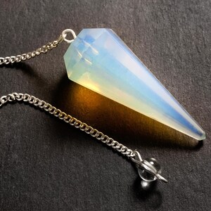 Könnte beinhalten: Ein facettierter, kegelförmiger Opalith-Anhänger an einer silberfarbenen Metallkette. Der Stein hat ein durchscheinendes, milchig-weißes Aussehen mit subtilen blauen und gelben Lichtreflexen. Eine kleine, klare, kugelförmige Perle ist an der Kette befestigt.