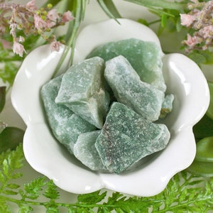 May include: A collection of rough, green aventurine stones fills a white, scalloped-edge bowl. The stones have a textured appearance and varying shades of green. The bowl is surrounded by greenery and small pink flowers, creating a natural aesthetic.