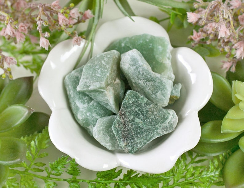 May include: A collection of rough, green aventurine stones fills a white, scalloped-edge bowl. The stones have a textured appearance and varying shades of green. The bowl is surrounded by greenery and small pink flowers, creating a natural aesthetic.