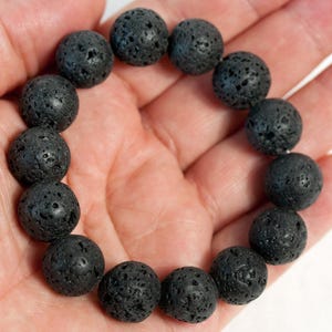 Peut inclure: Un bracelet en pierre de lave noire avec des perles rondes et poreuses. Le bracelet est composé de perles texturées de couleur gris foncé. Le bracelet est tenu dans une main, mettant en valeur les tons naturels et terreux de la roche volcanique.