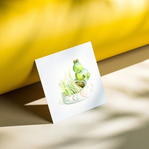 May include: A watercolor painting of a green frog sitting on the edge of a white planter with green grass growing inside. The planter has a white flower design.