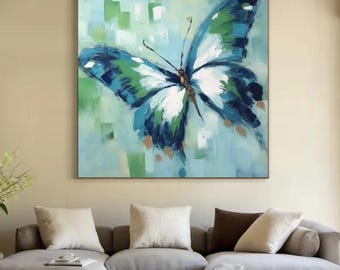 May include: Large square canvas art featuring a vibrant butterfly painting. The butterfly has blue, green, and white wings against a light blue and green background. The artwork is displayed above a modern gray sectional sofa.
