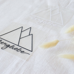 Könnte beinhalten: Zwei transparente Aufkleber mit einem Bergketten-Design und dem Text "bergliebe" in Schwarz und Weiß.