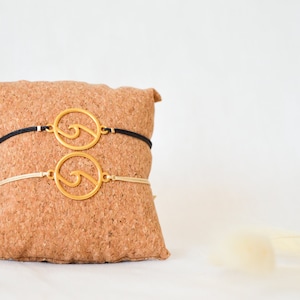 Könnte beinhalten: Zwei goldene Wellenarmbänder auf einem Kork-Display. Die Armbänder sind an schwarzen und beigefarbenen Schnüren.