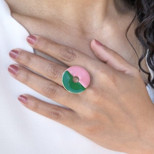 Könnte beinhalten: Ein Ring mit einem grünen und rosafarbenen Emaille-Design. Der Ring ist ein Kreis mit einem Loch in der Mitte. Der Ring befindet sich an einem Finger.