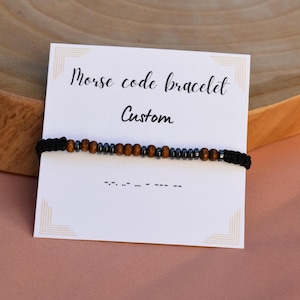 Peut inclure: Un bracelet en cordon noir avec des perles brunes et noires. Le bracelet est sur une carte blanche avec le texte "Bracelet code morse personnalisé" et le code morse du mot "Personnalisé".