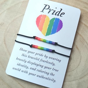 Peut inclure: Deux bracelets élastiques noirs avec des perles arc-en-ciel. Les bracelets sont sur une carte blanche avec un cœur arc-en-ciel et le texte "Pride" et "Montrez votre fierté en portant ce bracelet sans peur, en affichant courageusement votre véritable identité et en colorant le monde avec votre authenticité."