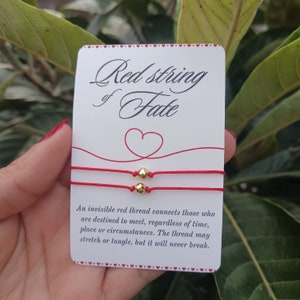 Könnte beinhalten: Zwei rote Armbänder mit goldenen Perlen. Die Karte lautet "Red string of Fate" und "Ein unsichtbarer roter Faden verbindet diejenigen, die dazu bestimmt sind, sich zu treffen, unabhängig von Zeit, Ort oder Umständen. Der Faden kann sich dehnen oder verheddern, aber er wird niemals reißen."