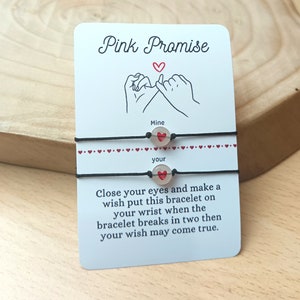 Puede incluir: Dos pulseras de amistad negras con dijes de corazón rojo. La tarjeta dice "Pink Promise" e incluye instrucciones para cerrar los ojos y pedir un deseo cuando la pulsera se rompa en dos.