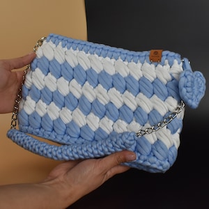 Puede incluir: Un bolso de mano de crochet azul claro y blanco con una correa de cadena plateada y un pequeño amuleto de crochet en forma de flor. El bolso tiene una textura tejida y una forma rectangular.