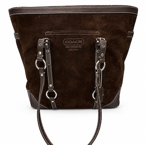 May include: A dark brown suede tote bag featuring smooth leather trim, silver-tone metal hardware, and dual shoulder straps. The front displays an embossed Coach Leatherware Est. 1941 logo. The bag includes decorative rings and a hanging leather tag.