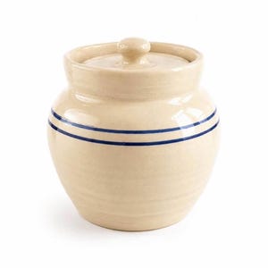 May include: A cream-colored ceramic crock with a matching lid and a rounded body. The exterior features two horizontal dark blue stripes encircling the center of the vessel. The surface has a smooth, glazed finish with a knob handle on the lid.