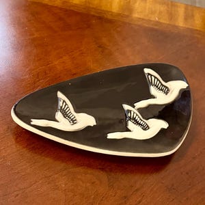 May include: A triangular ceramic trinket dish with a glossy black glaze featuring three raised white bird figures in flight. The dish has a smooth, curved edge and a white base rim, showcasing a high-contrast design with detailed wing patterns.