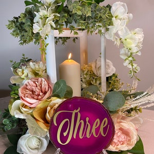 May include: A magenta agate slice table number with the word "Three" in gold script. The number is displayed on a clear stand, surrounded by a floral arrangement with a lit candle and a white lantern.