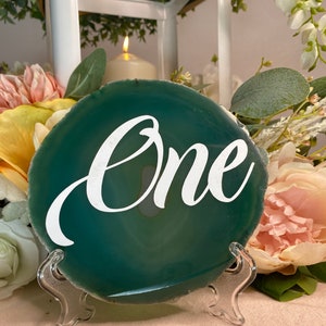 May include: Green agate slice table number holder with the word "One" written in white cursive lettering. The agate slice is on a clear plastic stand.