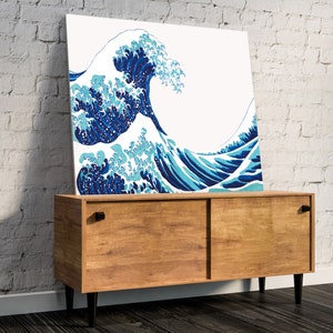 May include: A blue and white print of a large wave crashing against a white background. The print is framed and hanging on a wall above a wooden cabinet.