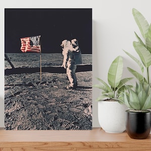 May include: A black and white photograph of an astronaut standing on the surface of the moon. The astronaut is wearing a white spacesuit and a backpack. An American flag is planted in the lunar soil behind the astronaut.