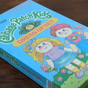 May include: A rectangular Cabbage Patch Kids Dress Up Set box featuring a light blue background with illustrations of two children, green grass, and flowers. The text Cabbage Patch Kids Dress Up Set is printed in green and red on the front cover.