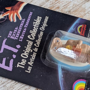 May include: A plastic figurine of E.T. the Extra-Terrestrial holding a small box. The figurine is packaged on a card with the text "E.T. The Original Collectibles" and "Les Articles de Collection Originaux".