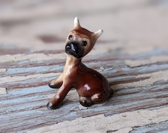 Può includere: Una piccola statuetta in ceramica di un boxer marrone e beige seduto con la testa alzata. La statuetta è dipinta con dettagli realistici, inclusi occhi neri e un naso nero.