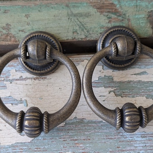 May include: Two antique bronze cabinet pulls with ring handles. Each pull has a circular backplate and a decorative ring handle. The pulls are made of metal and have a vintage, aged appearance. The background is a distressed, painted wooden surface.
