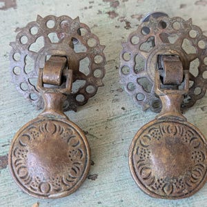 May include: Two antique bronze drawer pulls. Each pull features a decorative circular backplate with a floral design, a central handle, and a round, embossed front plate. The pulls have a vintage, aged appearance, suggesting they are vintage or antique.