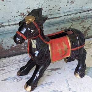 May include: A glossy black ceramic horse figurine featuring a red saddle with gold trim and a red bridle. The horse has gold-painted hooves and a gold accent on its forehead. The back of the horse has an open hollow compartment for storage.